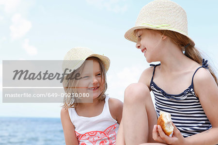 Two girls in sun hats eating sandwiches on beach, Scopello, Sicily, Italy