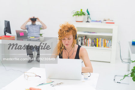 Businesswoman typing on laptop at office desk while listening to headphones