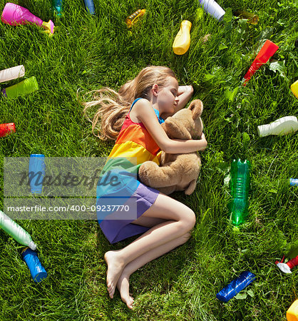 Young girl sleeping with her teddy bear in the plastic littered grass - pollution concept