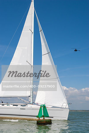 Sailing boat, sail boat or yacht with white sails passing a green navigation buoy at sea while a black helicopter flies over head