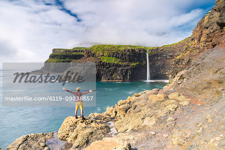 Man on cliffs with open arms admiring Gasadalur waterfall, Vagar island, Faroe Islands, Denmark, Europe