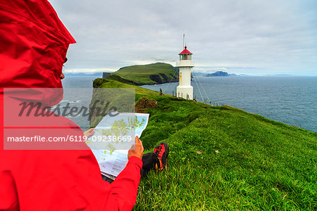 Hiker on cliffs looks at the map next to lighthouse, Mykines island, Faroe Islands, Denmark, Europe