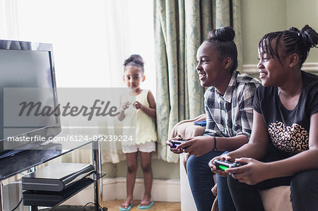 Toddler girl watching tween sisters playing video game in living room