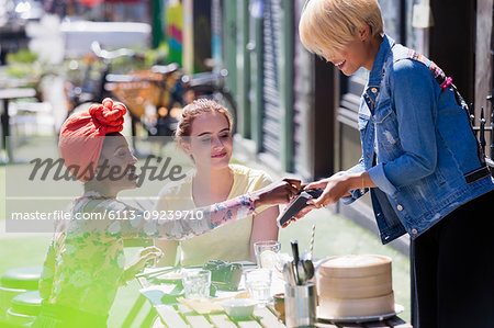 Young woman paying waitress with credit card at sunny sidewalk cafe