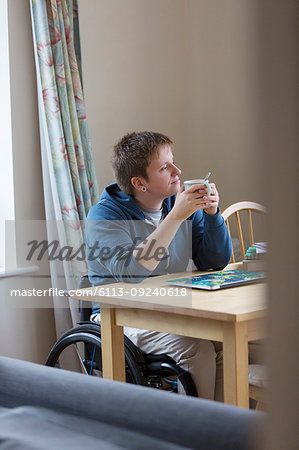 Thoughtful young woman in wheelchair drinking tea at dining table
