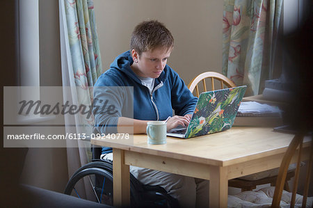 Focused young woman in wheelchair using laptop at dining table