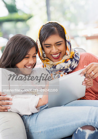Laughing, happy mother in hijab and daughter using digital tablet