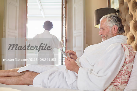 Mature man in spa bathrobe using digital tablet on hotel bed