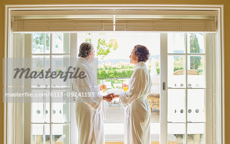 Mature couple in spa bathrobes drinking mimosas at hotel patio door