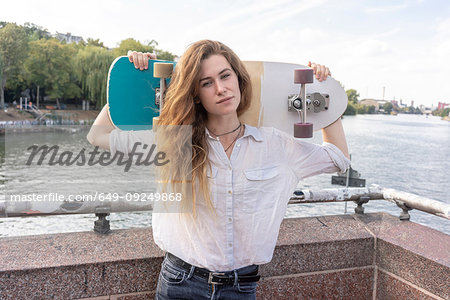Young woman with skateboard on shoulder, river in background, Berlin, Germany