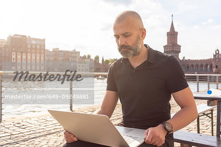 Man using laptop on bridge, river, Oberbaum bridge and buildings in background, Berlin, Germany