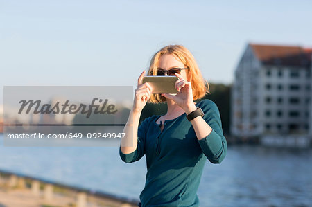 Young woman taking photograph with smartphone by river in summer, Berlin, Germany