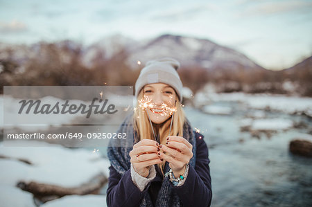 Woman in knitted hat holding sparklers on snow covered riverbank, portrait,  Orta, Piemonte, Italy