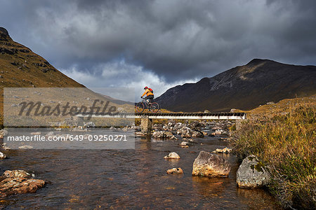 Male mountain biker biking over footbridge in mountain valley landscape,  Achnasheen, Scottish Highlands, Scotland