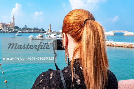 Female tourist with red hair photographing over sea toward Montaza palace, rear view, Alexandria, Egypt