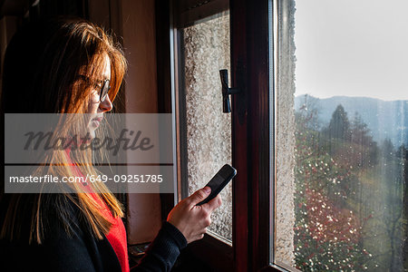 Young woman with long red hair looking at smartphone by window