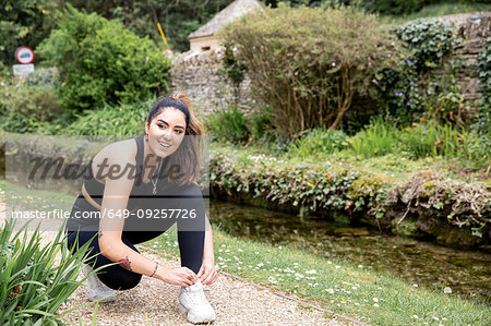 Young female runner tying trainer shoelaces on riverside path