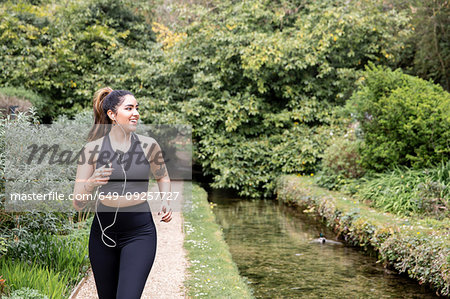 Young female runner listening to earphones while running on riverside path