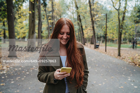 Young woman with long red hair looking at smartphone in tree lined autumn park, Florence, Tuscany, Italy