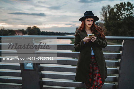 Young woman with long red hair leaning on footbridge looking at smartphone at dusk, Florence, Tuscany, Italy