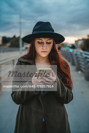 Young woman with long red hair on footbridge looking at smartphone at dusk