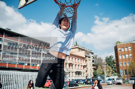 Young man hanging from basketball hoop at city basketball court