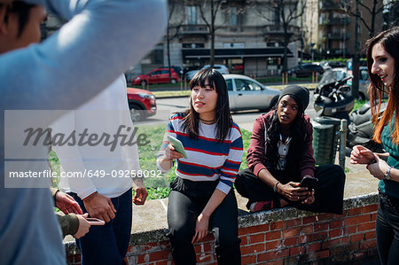 Young female and male adult friends sitting on wall chatting and looking at smartphones