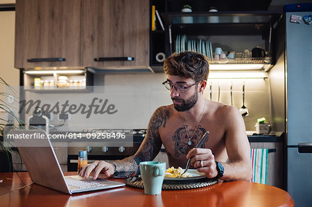 Mid adult man with tattoos eating omelette while typing on laptop