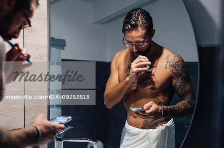 Mid adult man with tattoos brushing teeth and looking at smartphone at bathroom mirror, mirror image