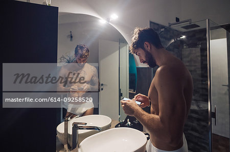 Mid adult man with tattoos using smartphone touchscreen at bathroom mirror, mirror image