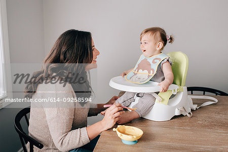 Mother feeding baby daughter in child seat on kitchen table