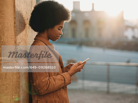 Young woman with afro hair using smartphone, leaning against stone wall, Florence, Toscana, Italy