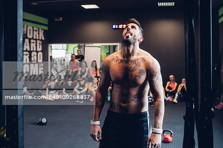 Young tattooed man training in gym, looking up at exercise bar