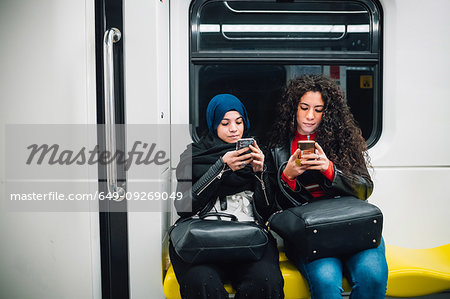 Young woman in hijab and friend sitting on subway train looking at smartphones