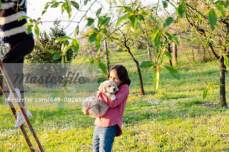 Girl holding a cute golden retriever puppy while her friend climbs tree ladder in orchard, Scandicci, Tuscany, Italy