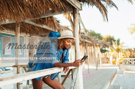 Young female backpacker using smart phone on beach hut patio
