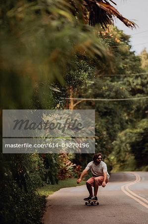 Mid adult male skateboarder crouching while skateboarding along rural road, Haiku, Hawaii, USA