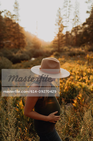 Mid adult pregnant woman in stetson with hands on stomach in rural valley at sunset, Mineral King, California, USA