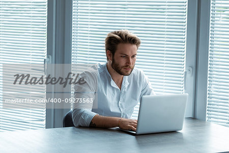 Serious young businessman working on a laptop computer with a look of concentration seated at an office table in front of large windows with blinds