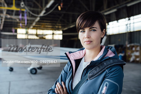 Confident young female pilot posing in the hangar with arms crossed, airplane on the background