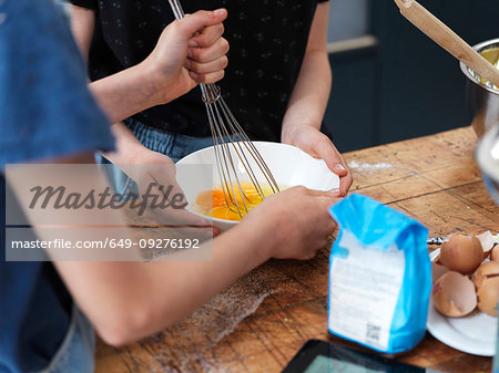 Girl and her sister baking a cake, whisking eggs in bowl on kitchen table, cropped view of hands