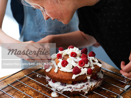Girl and her sister baking a cake, decorating cake with fresh cream and raspberries at kitchen table, cropped