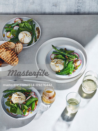 High key still life with glasses of white wine and plates of burrata spring salad on white table, overhead view