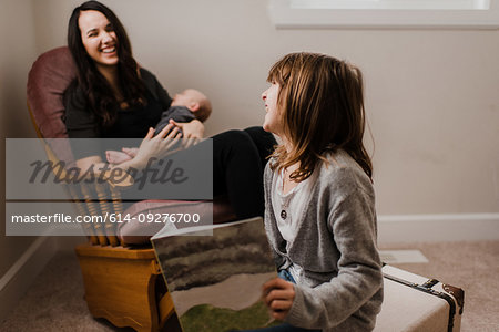 Girl laughing with mother while she cradles baby brother in living room armchair