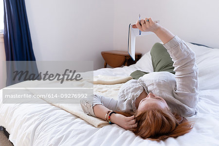 Young woman with red hair reclining on bed reading her diary