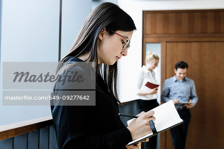 Young businesswoman writing notes in office, colleagues talking in background