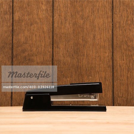 Black stapler on desk with wooden paneling.