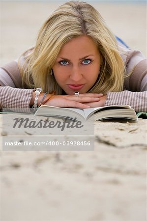 A beautiful blond haired blue eyed young woman laying on a beach reading her book