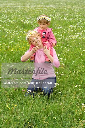 Baby girl sitting on her mothers neck wearing daisy wreath and picking more daisies on a meadow full of flowers