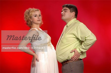 Kind of joke: who's belly is larger - pregnant woman's or her husband's?   Young couple on red background. Horizontal version.